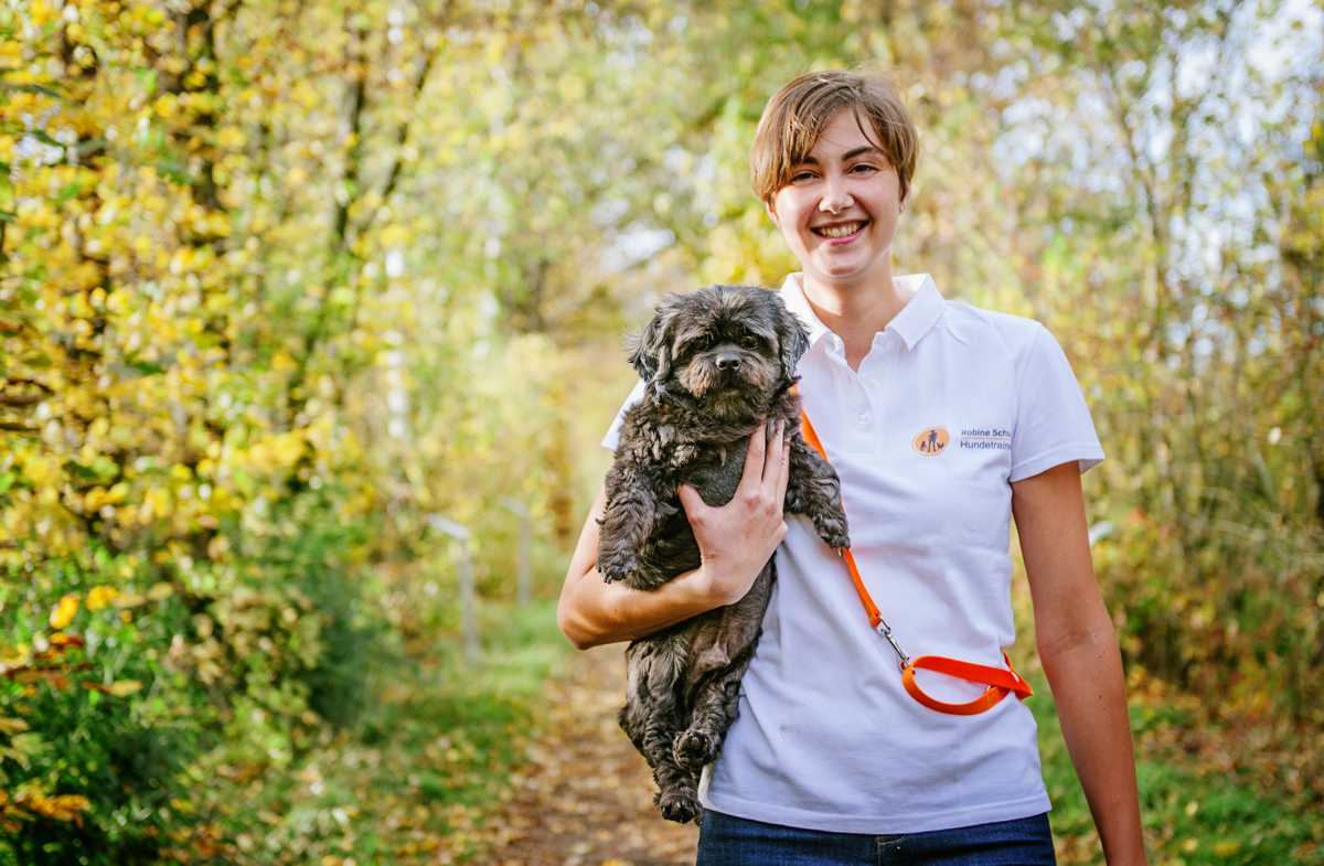 Robine Klinner trägt lachend einen kleinen Hund auf einem herbstlichen Waldweg